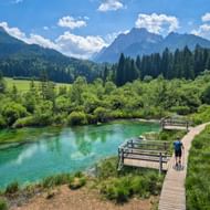 Houten looppad met uitkijkplatforms in natuurgebied Zelenci. Turquoise water omgeven door groene vegetatie, bos en bergen onder blauwe lucht.