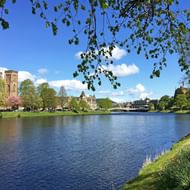 Rivier Ness door Inverness met historische stenen toren, brug en groene parken onder blauwe hemel met witte wolken.
