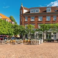 Geplaveide plein in Odense met café-terras, rode bakstenen gebouwen en groene bomen onder blauwe lucht.