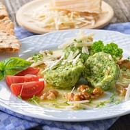 Twee groene kruidenknödels op wit bord met tomatenschijfjes, basilicum, peterselie, walnoten en kaas. Brood en kaaskommetje op achtergrond.