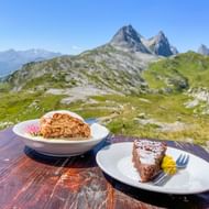 Twee gebakstukken op witte borden op een berghuttafel, met groene alpenweiden en rotstoppen van de Allgäu-Lechtal Alpen erachter.