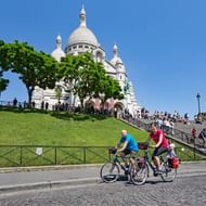 Twee fietsers in kleurrijke shirts rijden langs de witte Sacré-Cœur Basiliek in Parijs op een zonnige dag met toeristen op de trappen.