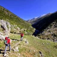 Twee wandelaars met rode rugzakken op de Hidden Valley trek in Albanië, lopend op een bergpad tussen groene hellingen en rotsachtig terrein.