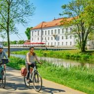 Twee fietsers op een verharde weg langs een kanaal in Oranienburg. Achter hen staat een wit barok kasteel met rood dak tussen groene bomen.