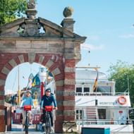 Twee fietsers met rode fietstassen rijden door een historische bakstenen poort in de haven van Emden, met boten en masten op de achtergrond.