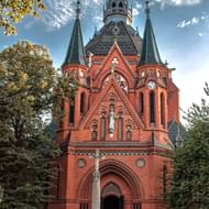 Neogotische rode bakstenen kerk in Břeclav met drie groene torenspitsen, gebogen ingangsportaal en stenen kruisbeeld ervoor, omlijst door bomen.
