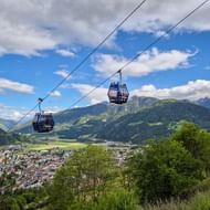 Twee blauwe gondels boven Sterzing met bergpanorama. De stad ligt in een vallei omringd door groene Alpentoppen onder een bewolkte hemel.