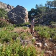 Wandelaar met gestrekte armen op rotsachtig pad op Rhodos, omringd door mediterrane vegetatie en rotsformaties onder heldere blauwe lucht.