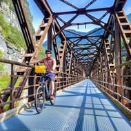 Fietser met helm en gele fietstassen steekt stalen vakwerkspoorwegbrug over in Chiusaforte, omgeven door groene bergen en bos.