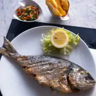Hele gegrilde vis op wit bord met schijfje citroen en slagarnituur, geserveerd met groentesalade en brood op marmeren tafel.