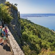 Drie wandelaars met rugzakken op stenen muur uitkijkend over Adriatische Zee en kust van Triëst vanuit Julische Alpen wandelpad.