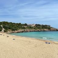 Zandstrand bij Portocolom met kristalhelder turquoise water, rotskliffen en mediterrane gebouwen op de heuvel onder blauwe lucht.