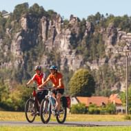 Twee fietsers in rode shirts op geasfalteerd fietspad met dramatische zandstenen kliffen van Saksisch Zwitserland op achtergrond en dorp.