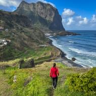 Persoon in rode jas lopend op grasrijk kustpad op Madeira met dramatische klif, oceaangolven en verspreide huizen op heuvel bij Porto da Cruz.