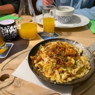 Gietijzeren pan met käsespätzle gegarneerd met gekarameliseerde uien en kruiden, geserveerd op houten plank met bierglazen en sinaasappelsap.