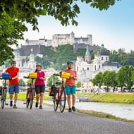 Drie fietsers met fietsen op pad langs de Salzach in Salzburg, met vesting Hohensalzburg en historische stadshorizon op achtergrond.