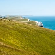 Glooiende groene heuvels lopen af naar witte krijtrotsen langs het turquoise water van Dorsets Jurassic Coast onder heldere hemel.