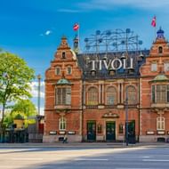 Rode bakstenen ingangsgebouw van Tivoli met sierlijke architectuur en groot verlicht bord. Deense vlaggen wapperen vanaf torens.