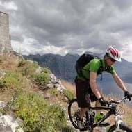Fietser in groen shirt en helm rijdt mountainbike op rotsachtig pad langs stenen ruïnes, met bergen en baai zichtbaar onder bewolkte lucht.