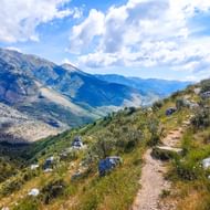 Kronkelend zandpad door Albanese bergen met rotsachtig terrein, schaarse vegetatie en dramatisch bergpanorama onder bewolkte hemel.