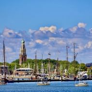 Haven van Enkhuizen met talrijke zeilboten en jachten afgemeerd in blauw water. Historische kerktoren met groene koepel rijst boven bomen uit.