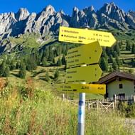 Gele wandelwegwijzer met richtingen naar Mandelwände bestemmingen met dramatische rotsachtige Alpentoppen en berghut op achtergrond.