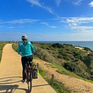 Vrouwelijke fietser in turquoise jas rijdend op verharde kustpad met uitzicht op Atlantische kliffen met groene vegetatie in Algarve.