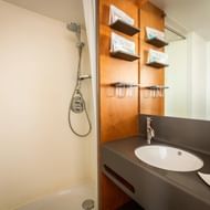 Compacte badkamer op MS Ave Maria met witte douche, ovale wastafel in donker blad, houten wandpanelen, planken en spiegel.