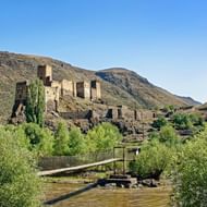 Khertvisi vesting ruïnes op een rotsachtige heuvel met stenen muren en torens. Een houten brug kruist een modderige rivier op de voorgrond.