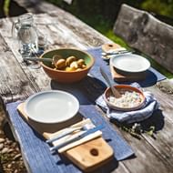Buitentafel met Bovec kaas in houten kom, olijven, lege borden, waterfles en snijplank op verweerd houten tafel.