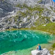Wandelaar met blauwe rugzak zit op rots bij turquoise alpenmeer in de Allgäuer Alpen, omringd door dramatische rotswanden en groene weiden.