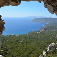 Panoramisch uitzicht op de kust van Rhodos door een natuurlijke stenen boog in Monolithos, met blauwe Middellandse Zee en beboste heuvels.