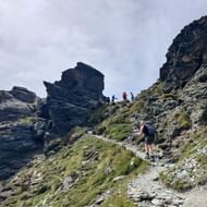 Wandelaars met rugzakken beklimmen rotsachtig pad naar Col de Riedmatten. Donkere rotsformaties en groene alpenvegetatie onder bewolkte hemel.