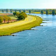 Luchtfoto van de Maas door groen landschap met gebogen oever, landbouwvelden en verspreide bomen onder blauwe hemel.