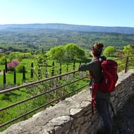 Wandelaar met rode rugzak op stenen terras met metalen leuning, uitkijkend over uitgestrekte groene Provence-vallei bij dorp Goult.