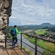 Wandelaar in groene jas met blauwe rugzak bij Bastei-uitkijkpunt met uitzicht op Elbe-vallei, Rathen en tafelbergen.