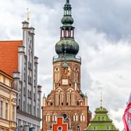 Gotische bakstenen kerktoren met groene koperen koepel in Greifswald, omringd door kleurrijke historische gebouwen met rode en groene gevels.