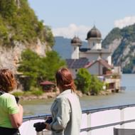 Twee vrouwen op een boot bekijken een Servisch klooster met koepeltorens bij de IJzeren Poort aan de Donau, omringd door steile kliffen.