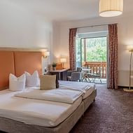 Ruime hotelkamer met tweepersoonsbed, oranje hoofdbord, zithoek en balkon met houten balustrade met uitzicht op groen bij Goiserer Mühle.