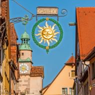 Sierlijk smeedijzeren Gasthof-bord met gouden zonnegelaat in groene krans, hangend boven straat in Rothenburg ob der Tauber met historische toren.