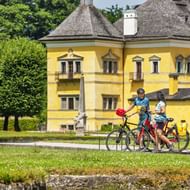 Twee fietsers met fietsen pauzeren voor het gele barokke kasteel Hellbrunn in Salzburg, met groene gazons en bomen zichtbaar.
