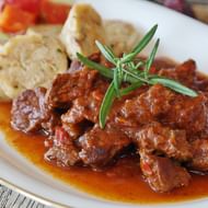 Bord met traditionele goulash met malse vlesstukken in rijke rode saus, gegarneerd met verse rozemarijn en geserveerd met gesneden knoedels.