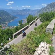 Twee fietsers op een kronkelende bergweg in Montenegro met panoramisch uitzicht op de baai van Kotor, bergen en kustplaats onder blauwe lucht.