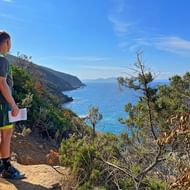 Wandelaar met rugzak en kaart op rotsachtig kustpad op Corsica, uitkijkend over blauwe Middellandse Zee met ruige kliffen.