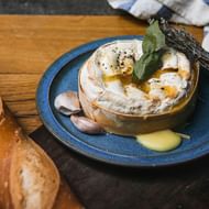 Gebakken Camembert kaas op blauw bord met laurierblaadjes, knoflookteentjes en kruiden. Vers brood en gestreepte keukendoek zichtbaar.