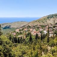 Luchtfoto van het dorp Qeparo genesteld in heuvels boven de Albanese kust, met traditionele huizen tussen groene vegetatie en blauwe zee.