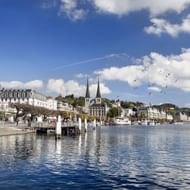 Lucerne's meer promenade met historische witte gebouwen, dubbele kerktorens en kalm meerwater dat blauwe lucht en wolken weerkaatst.