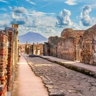 Oude Romeinse straat in Pompeii met ruïnes van bakstenen muren en zuilen. Vesuvius zichtbaar in de verte onder blauwe lucht met wolken.