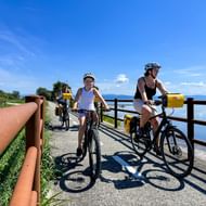Familie fietsers met gele fietstassen op Grado Lagune pad met houten hekken, uitzicht op blauw water en bergen in de verte.