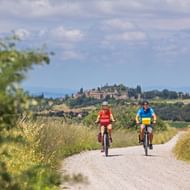 Twee fietsers op een grindpad door gouden velden in Toscane Val d'Orcia, met een bergdorp en cipressen op de achtergrond.